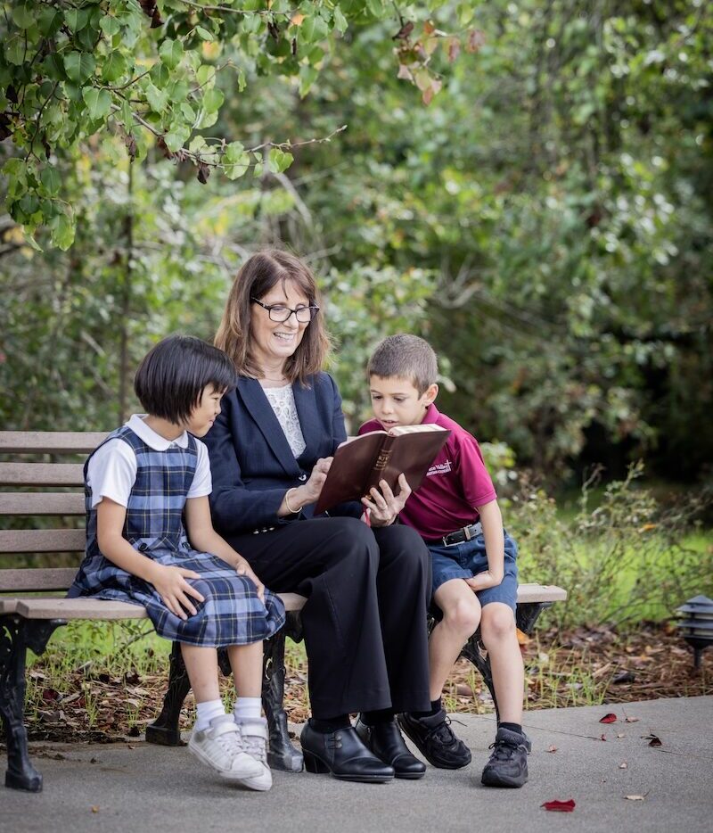 A teacher sits on a bench outdoors reading a book with two elementary students, creating a calm and engaging learning moment.