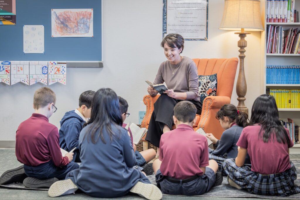 An elementary classroom scene where a teacher reads from a book while a small group of students in school uniforms sit in a circle on the carpet, following along in their own books. Bookshelves, student artwork, and a classroom calendar are visible in the background.