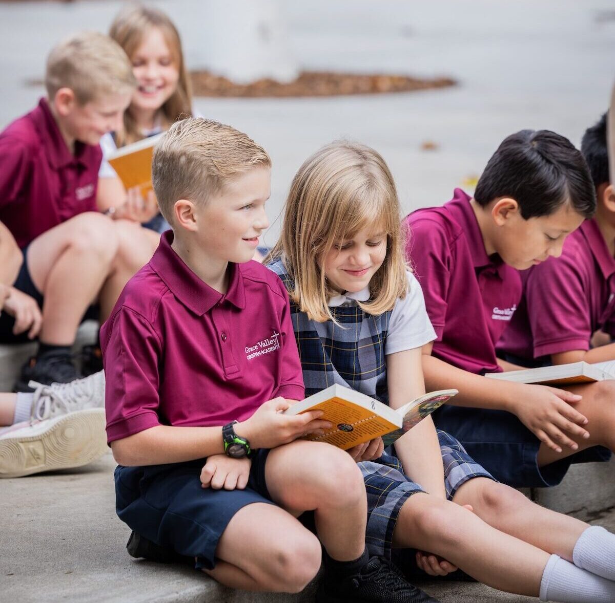 Fourth graders read literature outside. Two students share a book happily.