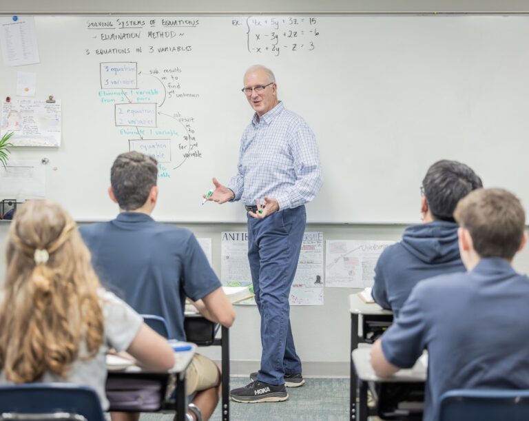 High School Algebra teacher explains solving a system of equations to his high school class. The white board behind him shows the problem they are working through.