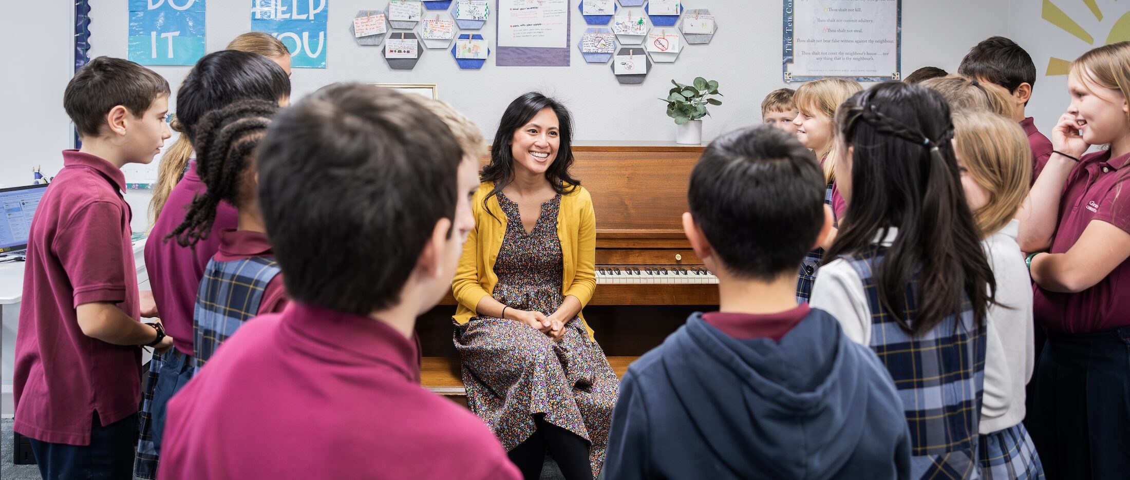 A teacher smiles while seated at a piano in the classroom, surrounded by attentive students.