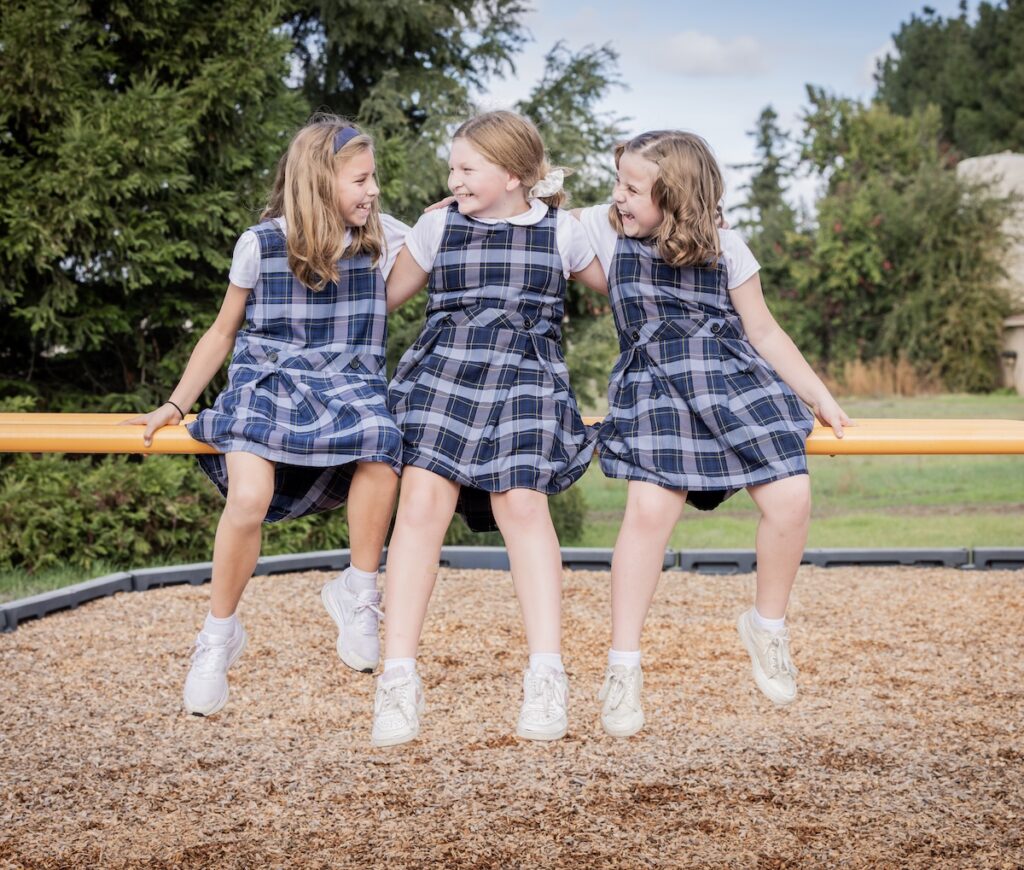 Three elementary aged girls sit on the play structure happily giggling with each other.