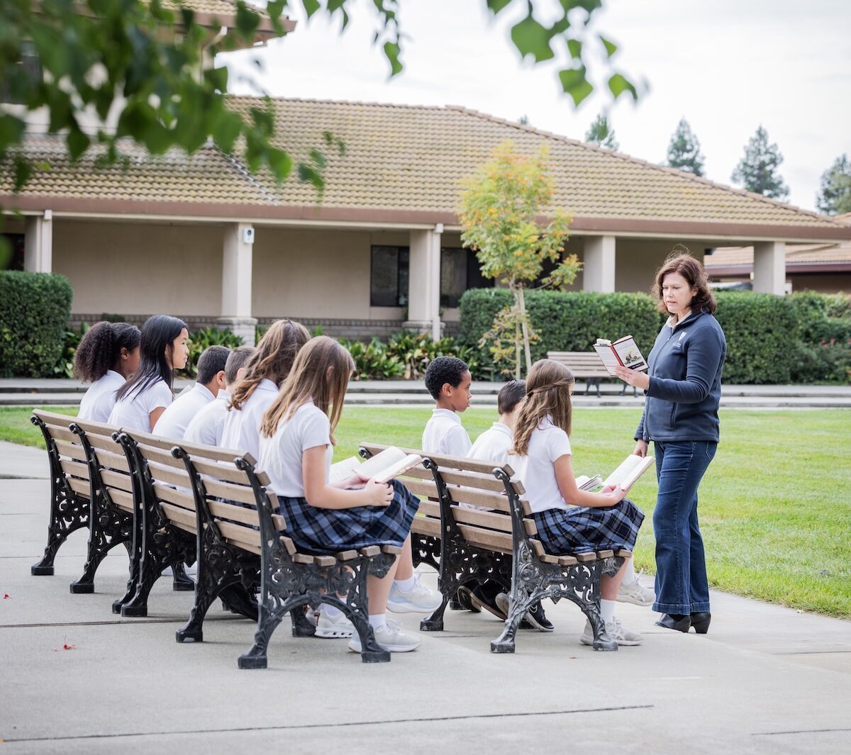 A Junior High class sits outside for literature class. The school building is in the background.