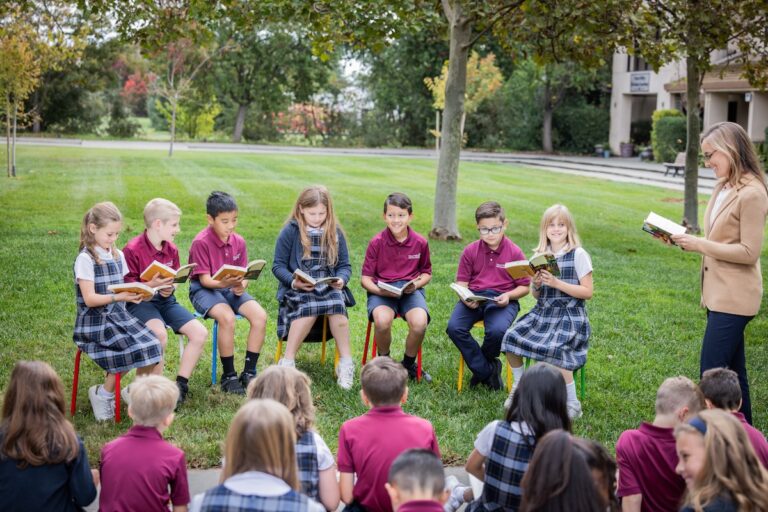 Some elementary students sit outside reading characters during literature class, as the teacher and rest of the class watch on.
