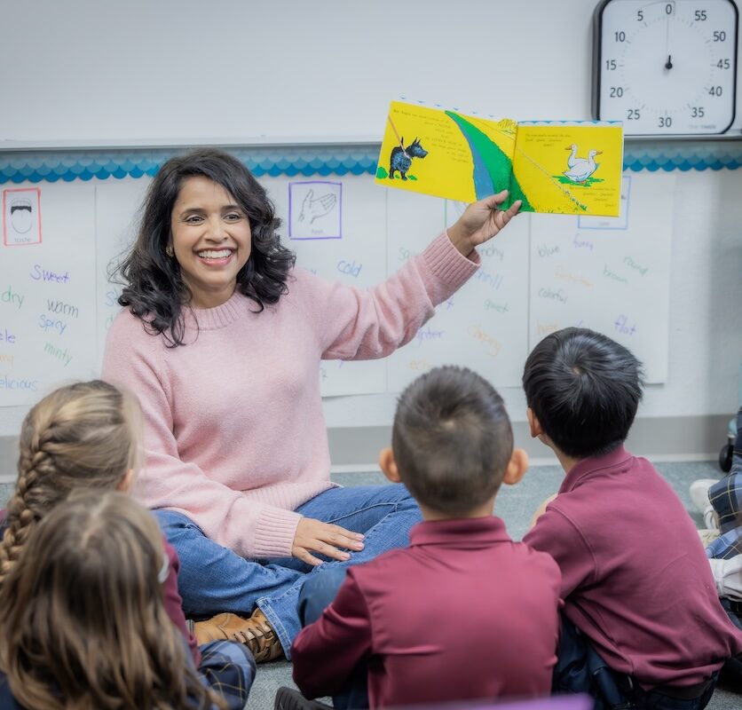 Principal Broderick sits on the floor with elementary students. She is animatedly reading a storybook to them.