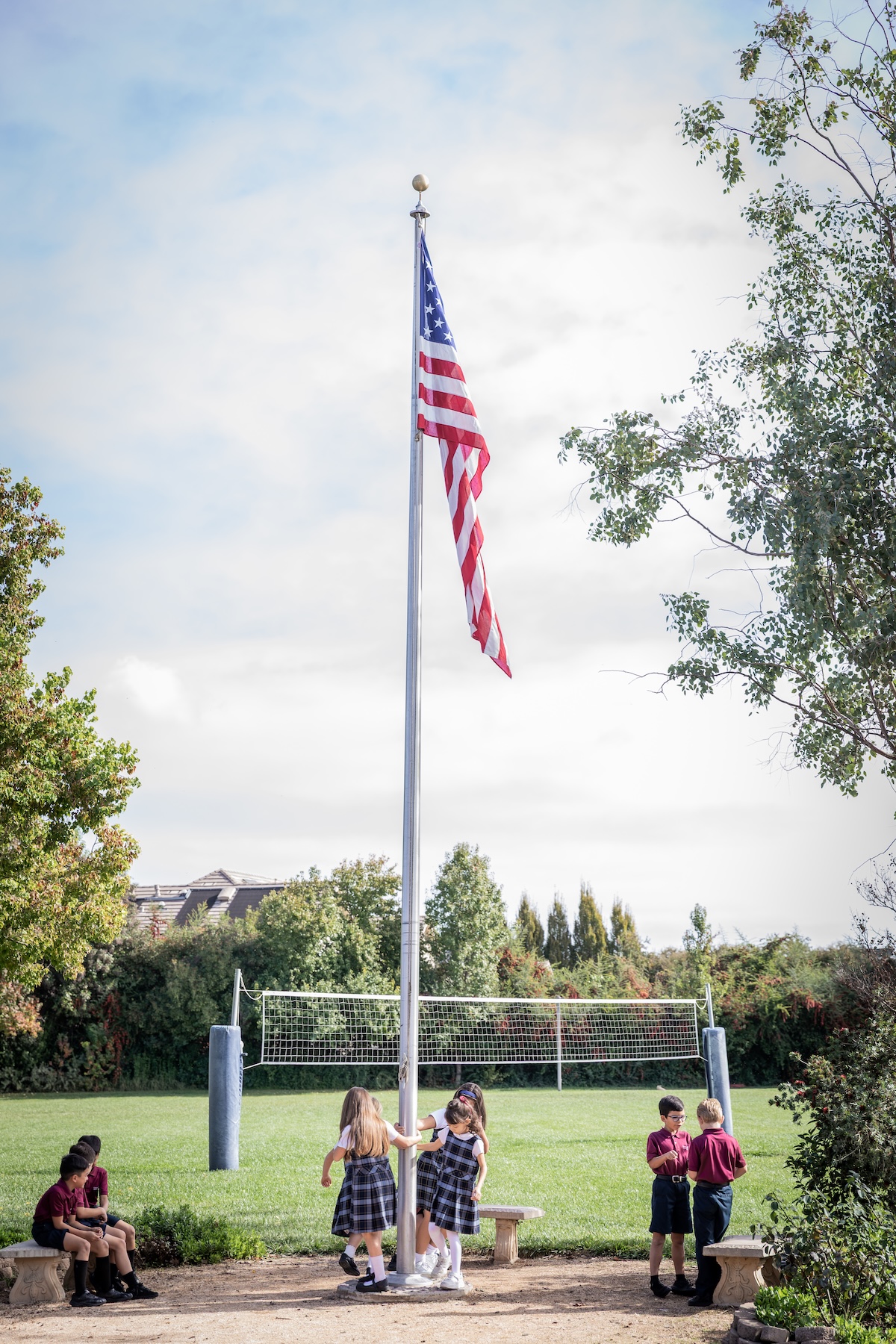 Elementary students play around a flagpole, with the American flag blowing gently in the wind.