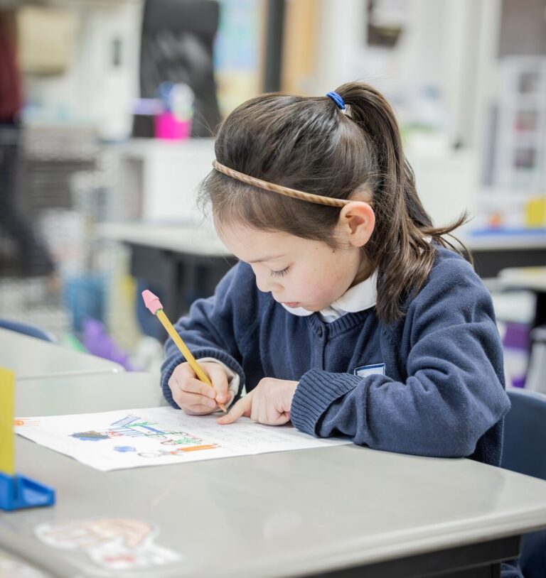 A kindergarten student writes a story at her desk to accompany her colorfully detailed drawing.