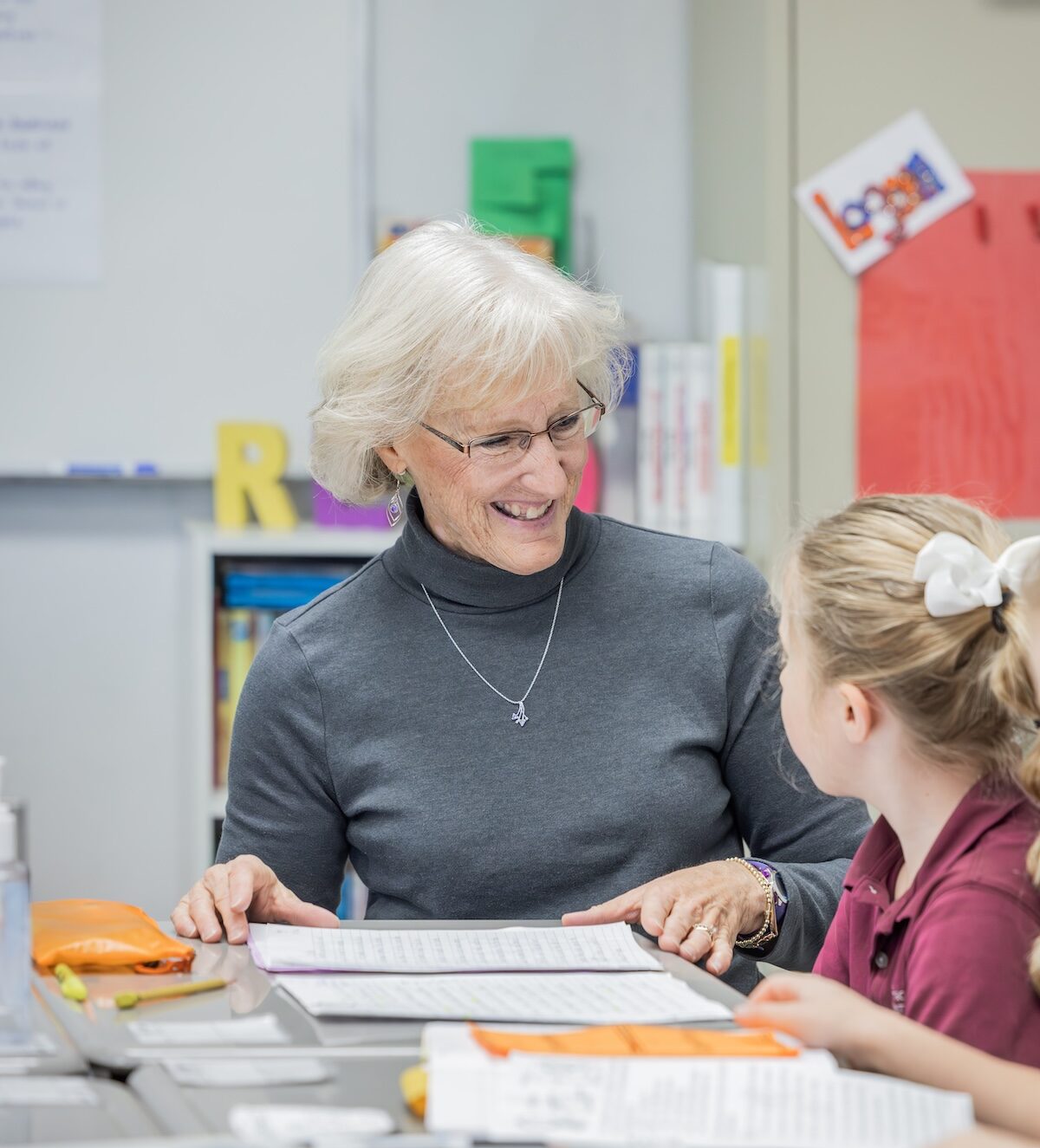 A teacher kneels at eye-level with a student, discussing the writing sample on her desk.
