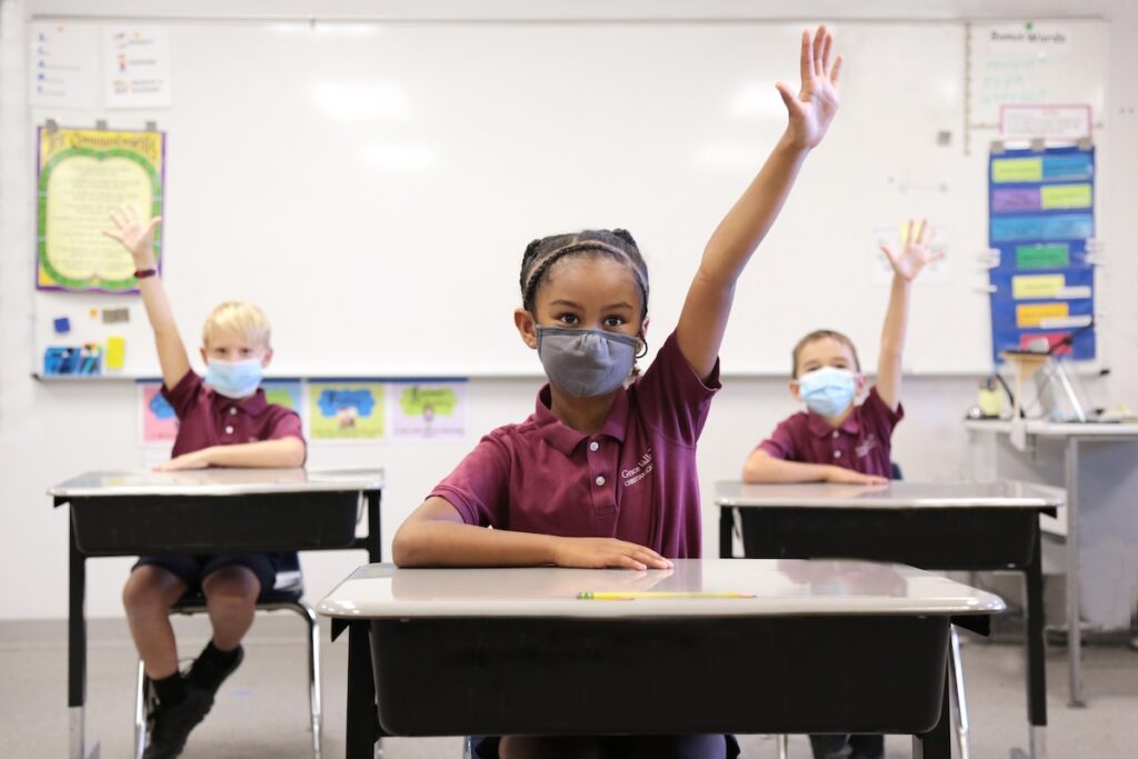Students excitedly raise their hands in class while wearing masks during COVID class instruction.